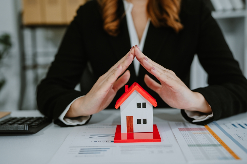 Hands forming a protective gesture over a model house to represent asset protection